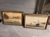 Pair of two signed Japanese paintings in ornate wooden gold-accented frames, resting on tiled floor against wall.