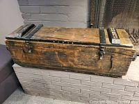 View of the vintage wooden ammunition crate closed, showing weathered wood, metal reinforcements, latch, and carved wooden blocks on the lid.