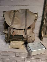 Front view of vintage military waterproof rucksack with leather harness straps, field dressing pouch, and metal field mess kit on a brick step