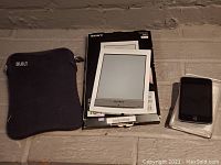 Sony Reader, its original box, protective carrying cover, USB cable, and Apple iPod Touch placed on table.