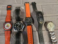 Five assorted wristwatches lying side by side on a gray textured surface.