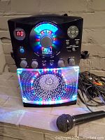 Front view of the Singing Machine karaoke machine powered on with multicolor lights and volume meter display, microphone connected and cables visible.