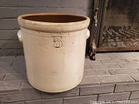 Antique five-gallon crock with cream exterior, brown interior, and number '5' marking, photographed from front and side views on a brick floor.