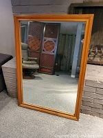 Front view of the large rectangular dresser mirror with a solid wood frame standing on the floor against a gray brick wall and a carpeted floor.