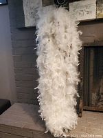 Full view of the white feather boa hanging vertically, showing the dense, fluffy texture and length of the boa against a brick and fireplace background.