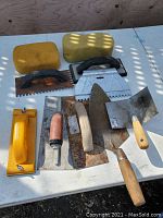 Photo showing assorted flooring and trowel tools laid on a white table including sponges, notched trowels, floats, and various hand trowels.