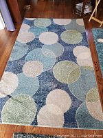 Full view of rectangular carpet with circular pattern in shades of blue, green, cream and beige on wooden floor. Wrinkle visible on lower left corner.
