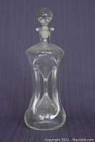 Clear vintage glass decanter upright with stopper in place, showing hourglass shape and round stopper.