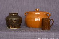 Three-piece antique ceramic lot with a small dark pot, a large orange cooking pot with lid and handles, and a small brown glazed cream jug, displayed against a grey fabric background.