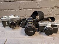 Three cameras side by side showing Olympus OM-10, Mamiya Sekor 500TL, and Olympus Trip 35