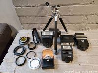 Full view of vintage camera accessories including two tripods, five flashes, and several lens filters with cases laid out on a surface.