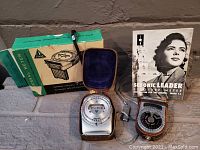Two vintage camera exposure meters displayed with their original leather cases, one with its original box and one with an instruction manual.