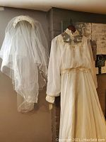 Full view of wedding dress and veil hanging side by side on wall.
