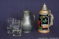 Group photo of lot showing pewter jug, German beer stein with lid, and two Bailey's promotional tumblers on purple cloth background.
