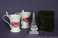 Photo showing two white ceramic teacups with pink rose floral patterns along with matching ceramic spoons, an antique lacquered green and red box with lid, and a silver pointed metal glasses holder.