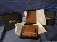 Two new Dante wallets displayed with their black boxes. One wallet is a brown leather bi-fold, the other a smaller brown leather wallet with a zipper pocket on the back.