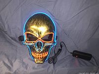 Gold skull mask lit up with blue outline and orange illuminated eye sockets and mouth, wired to battery pack
