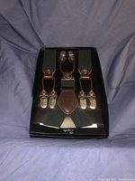 New set of black Zilpa suspenders displayed on black packaging box, showing three silver metal clips and brown leather connectors on black elastic fabric straps.