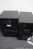 Two black metal filing cabinets side by side, one marked with writing and missing handle on top drawer, both showing signs of wear and rust.