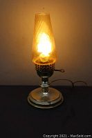 Small electric table lamp lit up, showing the metal base and clear glass chimney with swirling pattern.