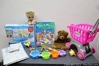 Overview of all toys including pottery wheel kit box, Groot plush, teddy bear, Munsch Mania board game, a pink toy shopping cart, and colorful fish-shaped sand toys on table