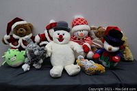 A group shot showing the collection of eight holiday themed and assorted stuffed toys including teddy bears, snowmen, a cat, a duck, and a green dragon/monster.