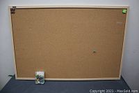 Front view of rectangular cork bulletin board with wooden frame and push pins on corners, standing on floor against wall.