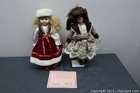 Two bisque porcelain dolls displayed standing side by side on a flat surface, one with blonde hair in red and white dress, the other with dark hair in floral dress and brown hat. Included is a certificate of authenticity.