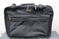 Black American Tourister suitcase front view showing two zippered front compartments and handle at top.