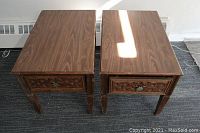 Two rectangular wooden end tables with wood grain tops and carved drawers next to each other on carpet.