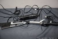 Three hair appliances laid out on a surface: a flat iron, curling iron, and hair dryer, all with cords.