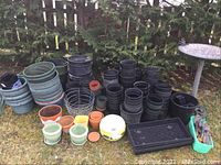 Wide view of many gardening pots stacked in groups of different sizes and colors, arranged on grass with a wooden fence and shrubbery in background.