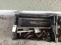 Plastic crate containing various metal hand tools like C-clamps, pipe clamps, hand clamps, and drill bits in a small tray within the crate.