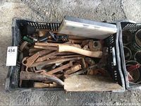 Top view of a black plastic crate filled with rusty hand tools including axes, saws, and large clamps.