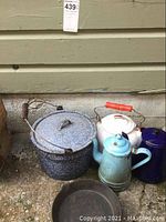 Photo showing enamel pot with lid, white and blue enamel kettles grouped together on ground near a wall.