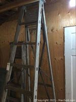 Front view of an 8-foot wooden A-frame ladder leaning against a wooden wall indoors showing multiple wear marks and wood grain.