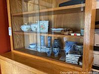 Wide shot showing wooden cabinet with multiple shelves of ceramic and decorative items including covered dishes, pitchers, cups, vases, tiles, and wooden pieces.