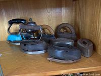 Shelf display showing six antique clothes irons made of cast iron, mostly charcoal type, plus one vintage electric iron with a black handle and blue body.