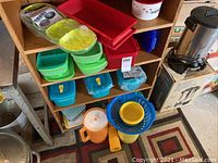 Multiple colorful Tupperware containers on wooden shelves and floor, including green, blue, red containers and a yellow pitcher.