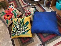 Four decorative cushions including three embroidered floral needlepoint cushions and one solid blue cushion on patterned rug.