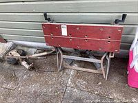 Photo showing folding wooden workbench with red top and metal legs folded, placed outdoors on concrete near wall.