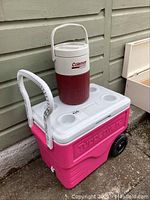 Pink Igloo wheeled cooler with white lid and handle, holding a Coleman red and white portable drink cooler jug.