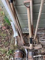 Photo showing six weathered garden tools leaning against a house exterior wall including two shovels, a cultivator, a hoe, a garden fork and a rusty metal container on the ground.
