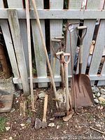Photo of garden tools including shovel, hoes, rake and axes leaning against a fence, showing weathered and rusty condition.