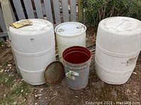 Full view of four barrels outdoors on ground near fence and foliage. Two large white barrels, one smaller white barrel, one metal barrel with a lid.