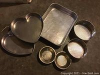 Various metal baking pans including a heart shaped pan, rectangular pan, and four round pans with perforated or mesh bottoms on a dark counter.