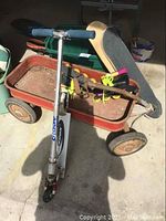 Photo showing the red metal wagon loaded with a silver Blade scooter with blue handlebars, rollerblades with neon yellow laces, and skateboards inside the wagon.