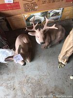 Photo showing both cement deer statues, the buck with antlers and the doe lying down, placed on a concrete surface near cardboard boxes.