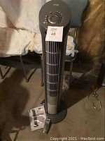 Full view of the tower fan standing on the floor next to chairs, control panel on top, instruction manual and remote visible on the floor.