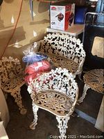 White painted cast iron loveseat and chairs with vine motif and bag of cushions on bench